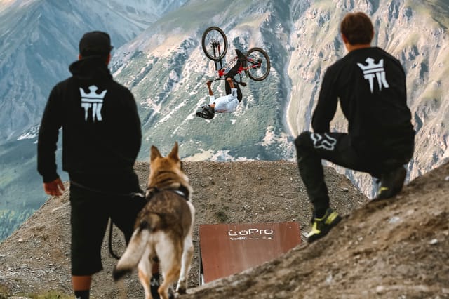 Friends and a dog watching a mountain biker backflip over the valley — Mottolino Bikepark, Livigno